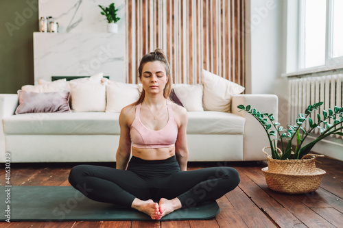 Attractive young woman sit in lotus yoga position at home in living room with her eyes closed. Relaxing state of mind, meditation.