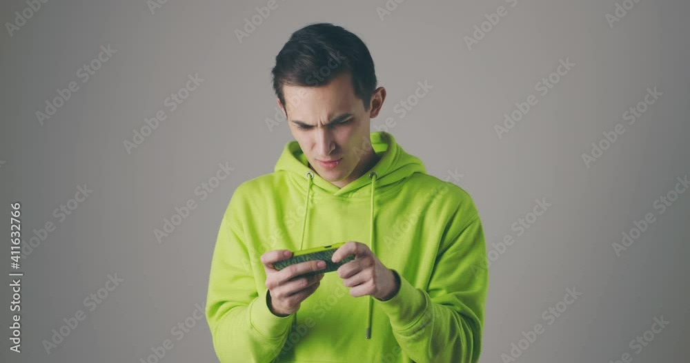 Handsome young man in green sweatshot playing game on smartphone at grey studio wall. Using modern technology - apps, social networks. Handsome young man in green sweatshot playing game on smartphone at grey studio wall. Using modern technology - apps, social networks.