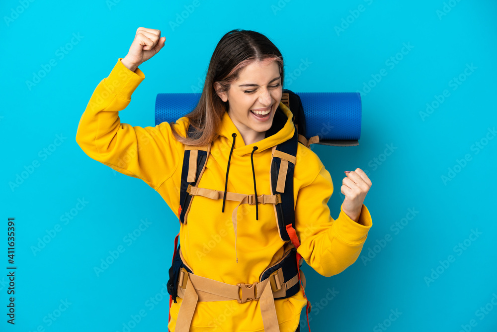 Young mountaineer woman with a big backpack isolated on blue background celebrating a victory Young mountaineer woman with a big backpack isolated on blue background celebrating a victory