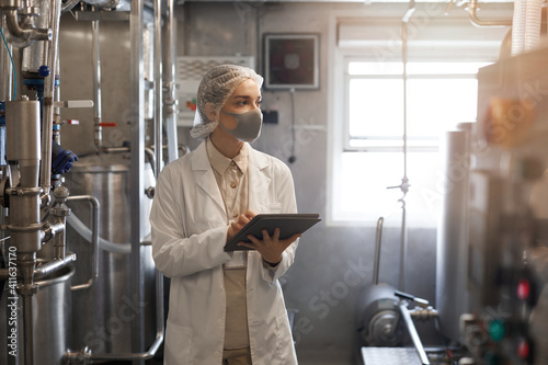 Waist up portrait of young woman wearing mask and holding digital tablet during quality control inspection in sunlit workshop at food factory, copy space photo