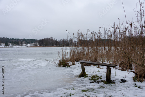 Fototapeta Naklejka Na Ścianę i Meble -  Old damaged wooden bench by the frozen lake. Typical polish winter landscape. Selective focus. Rzeck, Masuria, Poland. 