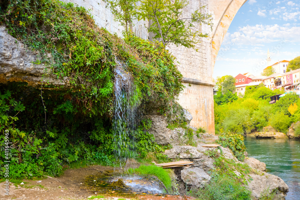 A small waterfall next to the historic Old Mostar Bridge or Stari Most with the village of Mostar, Bosnia and Herzegovina in view over the Neretva River.