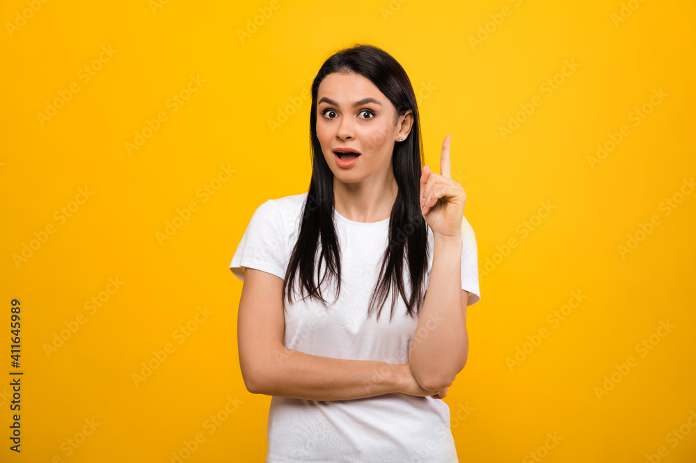 A pretty excited caucasian brunette woman in white basic t-shirt, got a new idea, standing on isolated orange background, looks directly at the camera A pretty excited caucasian brunette woman in white basic t-shirt, got a new idea, standing on isolated orange background, looks directly at the camera
