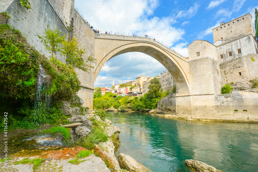 A small waterfall at the base of the Mostar Bridge in Bosnia and Herzegovina as tourists enjoy a view of the emerald green water of the river Neretva