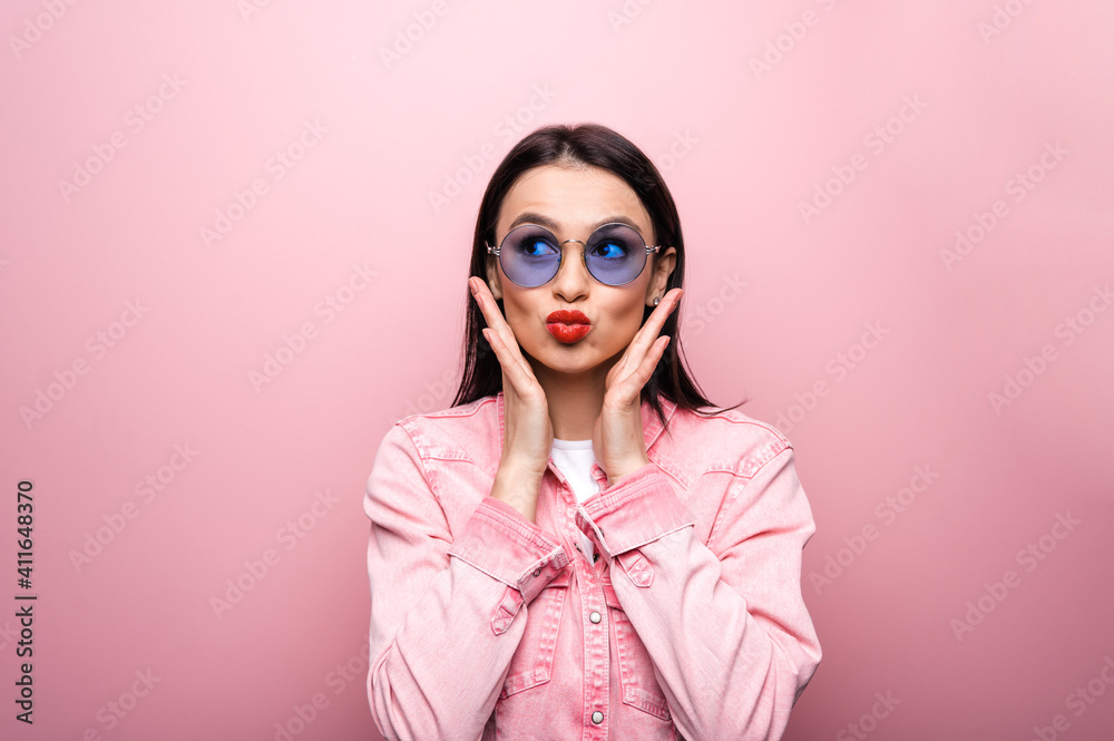 Attractive happy fashionable caucasian young woman in sunglasses, posing on isolated pastel pink background, shows kiss gesture