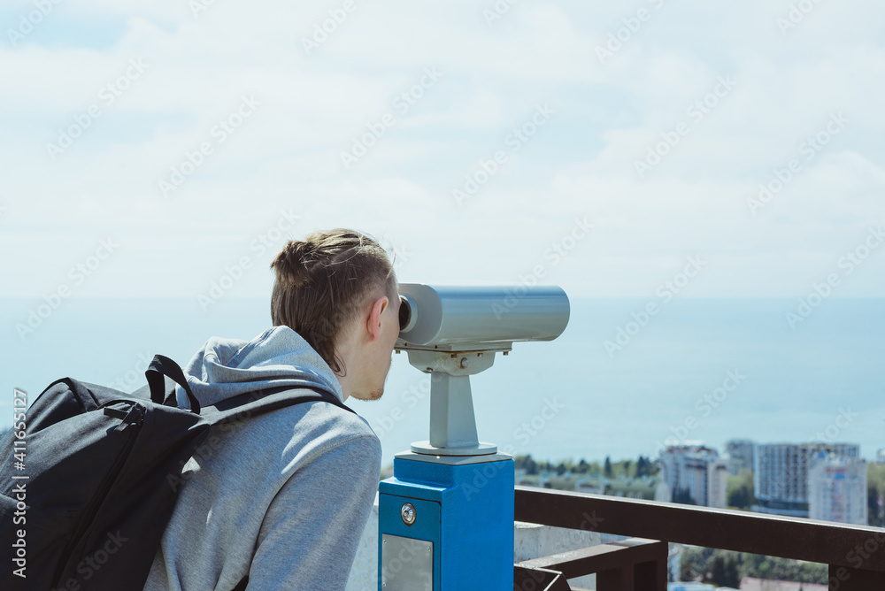 young hipster tourist man looking through metal coin operated binoculars on sea, sky and city, horizontal lifestyle stock photo image