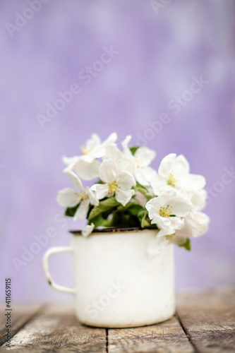 Flowers of apple trees on branch, collected in bouquet. Still life of an old iron mug and flowering branches of fruit tree on a purple blurred background