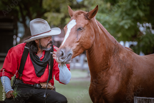 Portrait cowboy wearing dress vintage style and hat pose taking care and feed his brown horse by love and care.  © APchanel