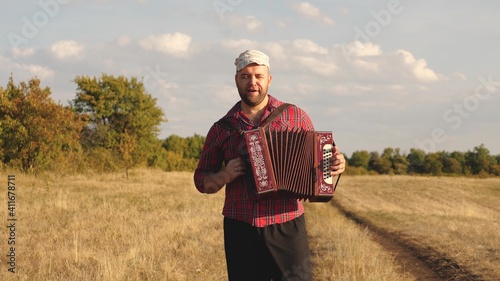 Man plays accordion in sun. Artist performs in open air, sings and plays musical instrument. Tradition and celebration in countryside. Cheerful man is a musician, he sings and plays musical instrument