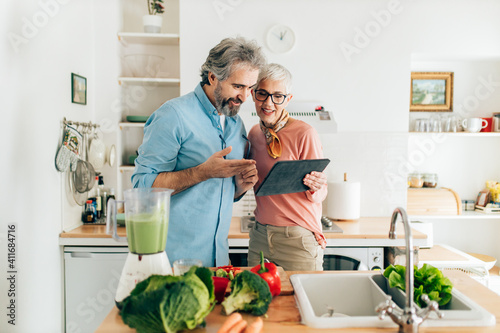 Senior couple preparing healthy smoothie in kitchen and using tablet to read recipe