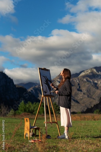Young beautiful girl paints a picture on an easel in the mountains. Artist profile with brush and paint palette. Inspiration by the autumn landscape of the Altai Mountains.