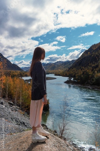 A young beautiful girl stands on a rock above the river in the mountains. © AngelinaProtein