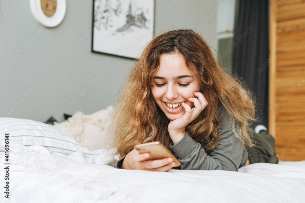 Beautiful smiling teenager girl with curly hair using mobile phone on bed in her room remote communication Beautiful smiling teenager girl with curly hair using mobile phone on bed in her room remote communication