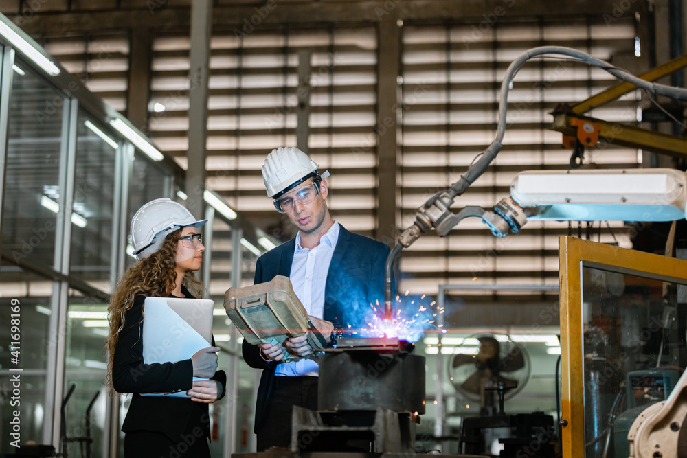 man engineer and female assistant wearing black suit and white hard hat in factory male engineering using automatic box controls robotic arms machine. industrial technology concept. man engineer and female assistant wearing black suit and white hard hat in factory male engineering using automatic box controls robotic arms machine. industrial technology concept.