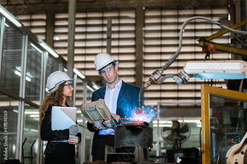 man engineer and female assistant wearing black suit and white hard hat in factory male engineering using automatic box controls robotic arms machine. industrial technology concept.