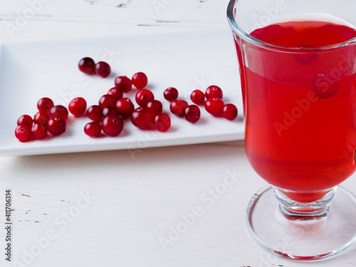 Selective focus on cranberries in a fresh drink in a glass cup on a white wooden background. Fresh ripe berries are scattered on a white rectangular ceramic plate. Copy space. 