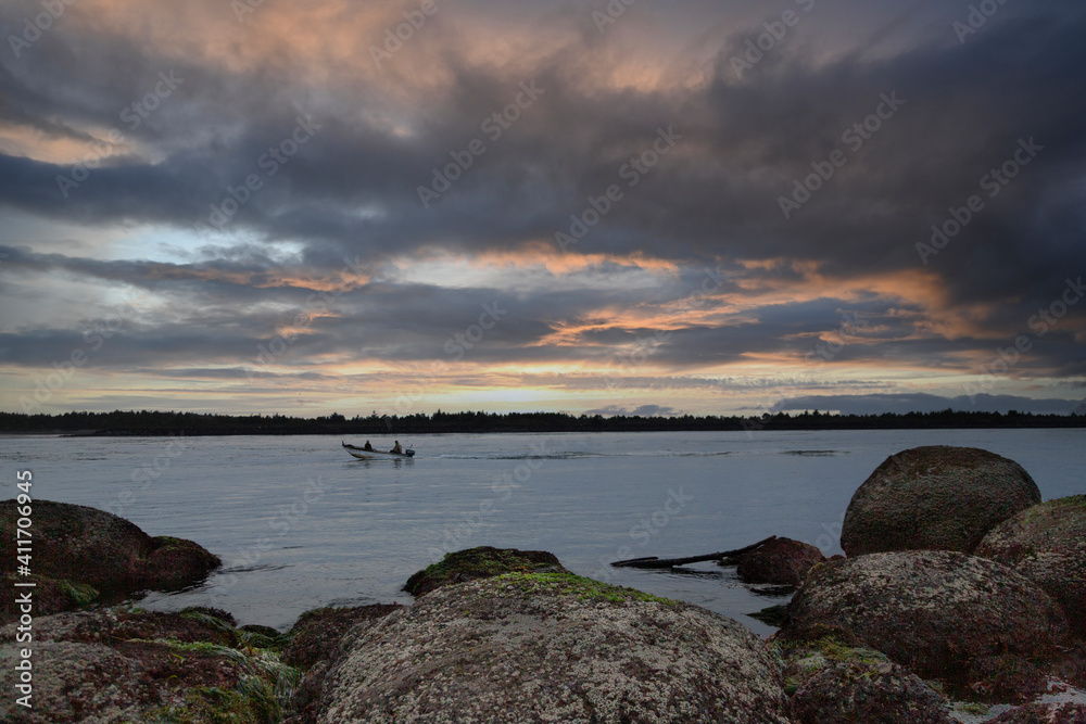 A wide angle view of a bay inlet at dusk with rocks in the foreground, a boat in the bay and dramatic, colorful clouds overhead 