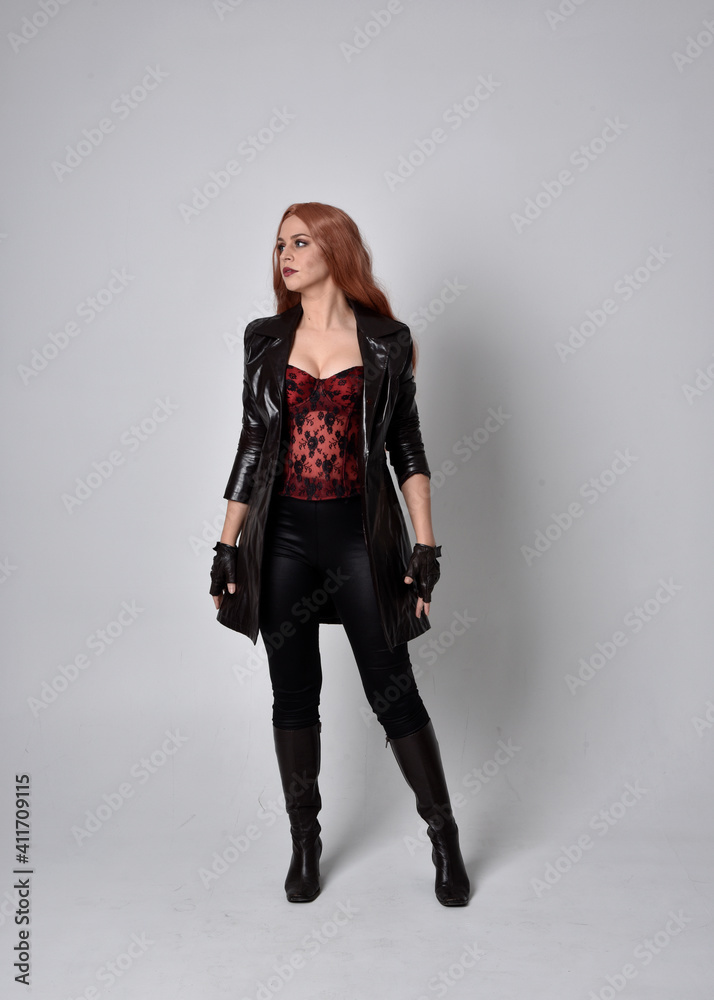 full length portrait of girl with long red hair wearing dark leather coat, corset and boots. Standing pose facing front on with magical hand gestures against a studio background. full length portrait of girl with long red hair wearing dark leather coat, corset and boots. Standing pose facing front on with magical hand gestures against a studio background.
