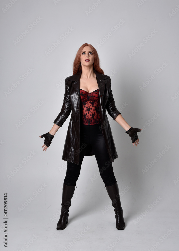 full length portrait of girl with long red hair wearing dark leather coat, corset and boots. Standing pose facing front on with magical hand gestures against a studio background. full length portrait of girl with long red hair wearing dark leather coat, corset and boots. Standing pose facing front on with magical hand gestures against a studio background.