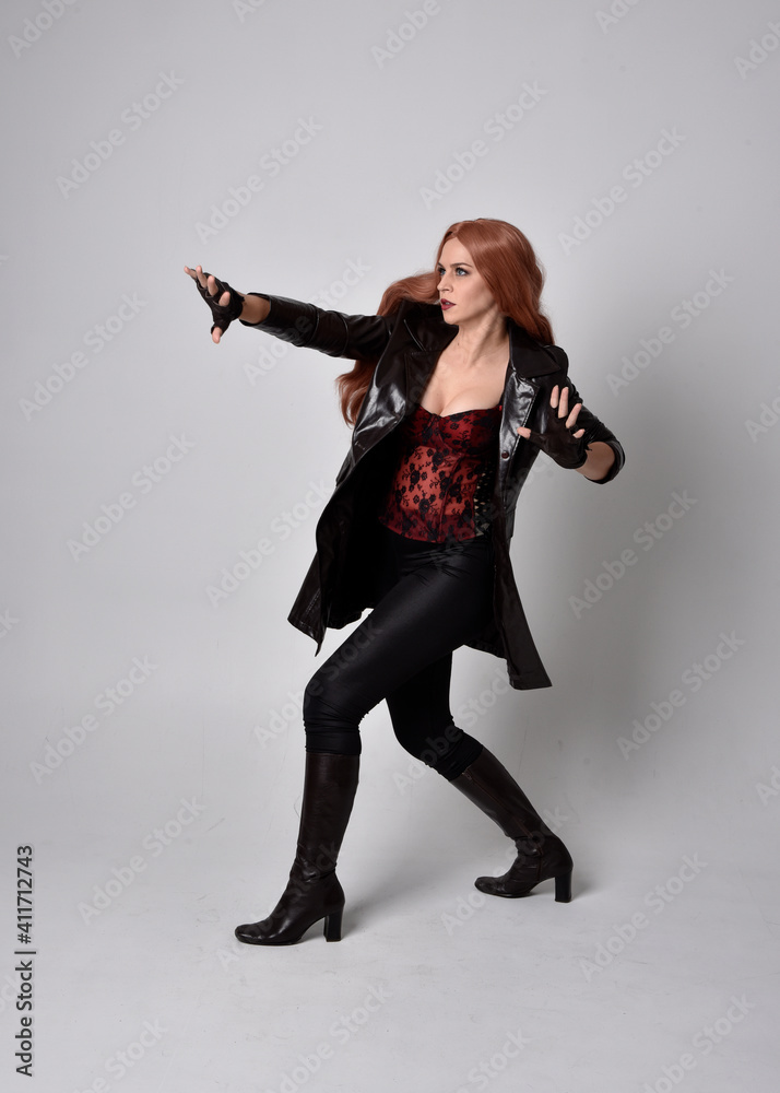 full length portrait of girl with long red hair wearing dark leather coat, corset and boots. Standing pose facing front on with  magical hand gestures against a  studio background.