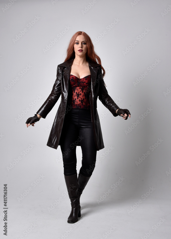 full length portrait of girl with long red hair wearing dark leather coat, corset and boots. Standing pose facing front on with magical hand gestures against a studio background. full length portrait of girl with long red hair wearing dark leather coat, corset and boots. Standing pose facing front on with magical hand gestures against a studio background.