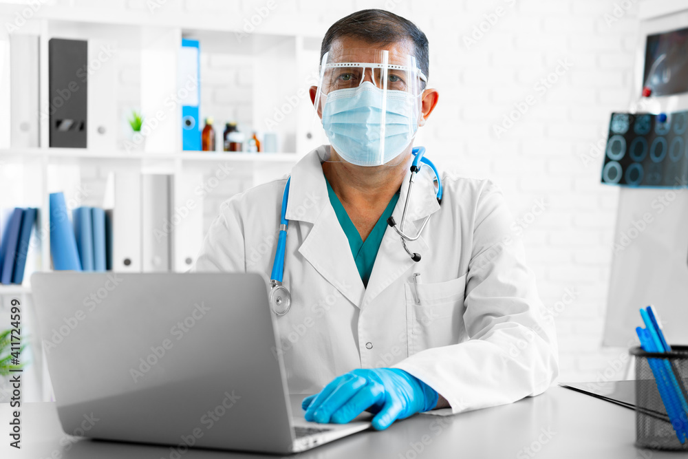 Middle-aged male doctor in face shield and mask sitting at the table at his office Middle-aged male doctor in face shield and mask sitting at the table at his office