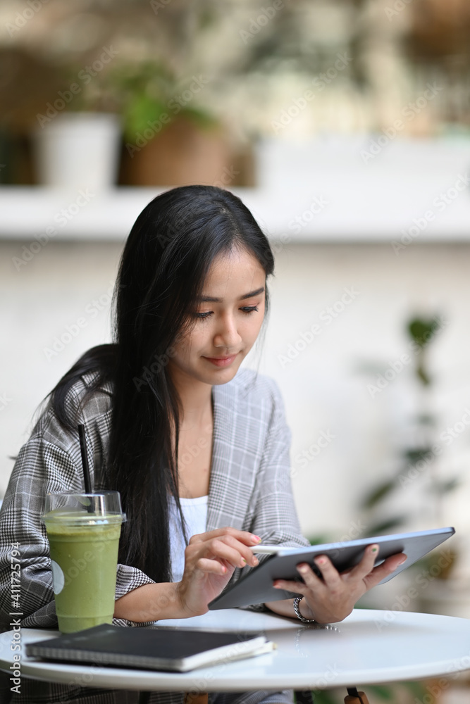 Portrait of young woman resting in cafe with green fresh vegetable juice and working on digital tablet.