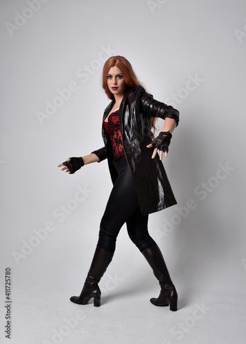 full length portrait of girl with long red hair wearing dark leather coat, corset and boots. Standing pose facing front on with magical hand gestures against a studio background, low camera angle.