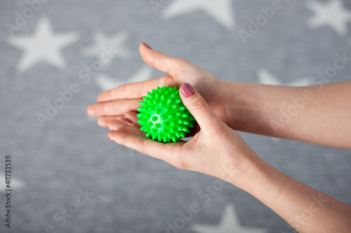 close-up two female palms hold a green spiky massage ball on a gray carpet with white stars.