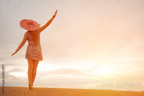 Fototapeta Naklejka Na Ścianę i Meble -  silhouette of a romantic and dreaming young woman walking down the slope of a sand dune with her hands up. Female silhouette in a hat and short dress against the backdrop of the sunset sky. copy space