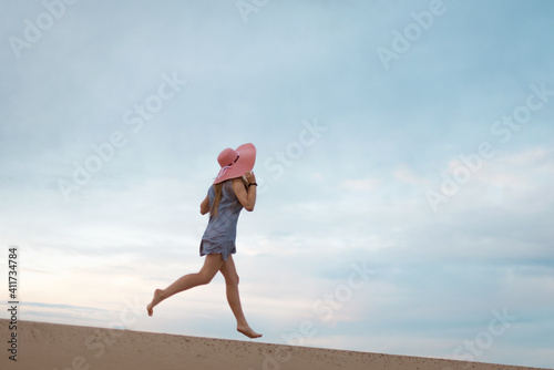 silhouette of a romantic and dreaming young woman running down the sand dune slope. Female silhouette in a hat and short dress against the backdrop of the sunset sky.