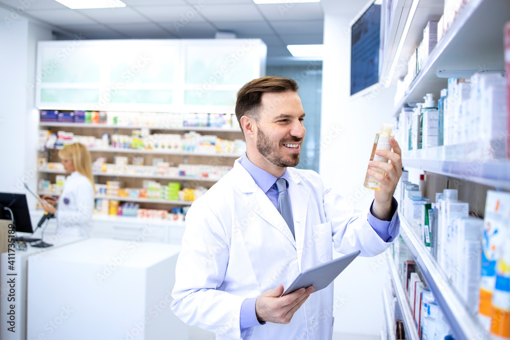 Handsome male pharmacist in white coat working in pharmacy store or drugstore. Checking medicines on his tablet computer. Healthcare and apothecary.