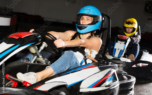 Young female in helmet and other people driving cars for racing in sport club indoor