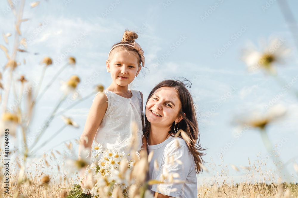 Beautiful young mother and her daughter having fun at the field. Chamomile on foreground, sky background. White clothes. Beautiful young mother and her daughter having fun at the field. Chamomile on foreground, sky background. White clothes.