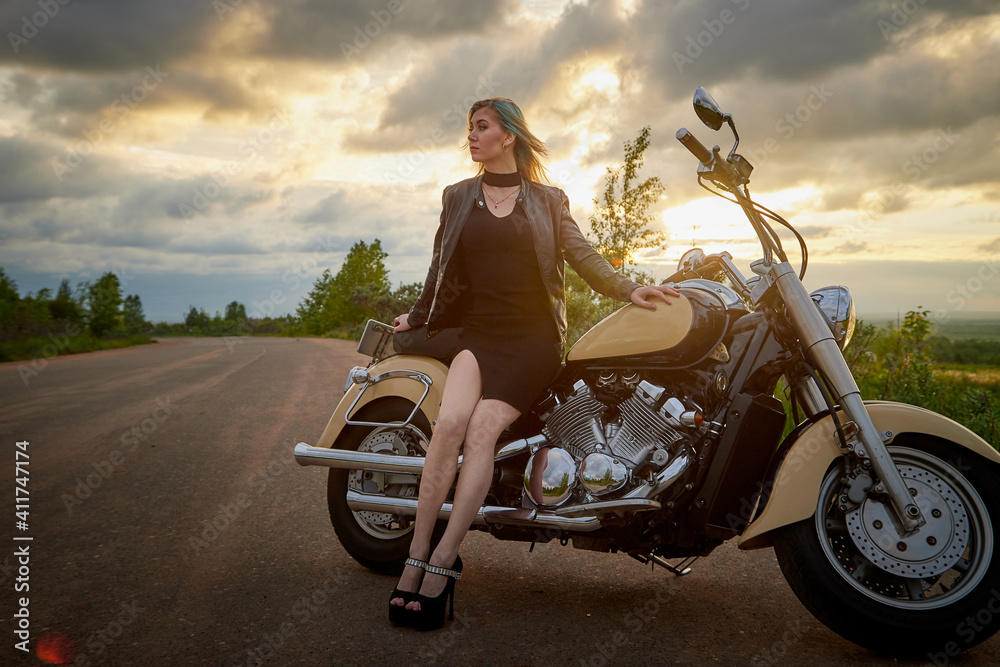 Fototapeta premium Attractive female brunette motorcyclist with motorcycle in a summer evening during sunset. Beautiful girl on nature and sky with sun and clouds background. Adventure concept