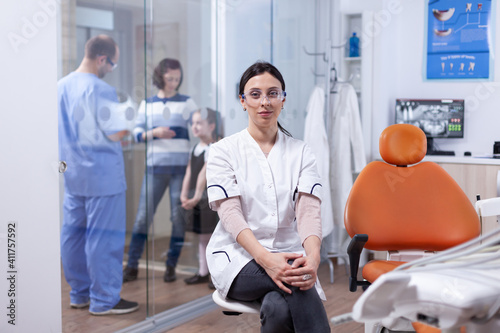Dentist in dentistiry office sitting on chair while assistant is talking with mother and her little girl. Stomatolog in professioanl teeth clinic smiling wearing uniform looking at camera.