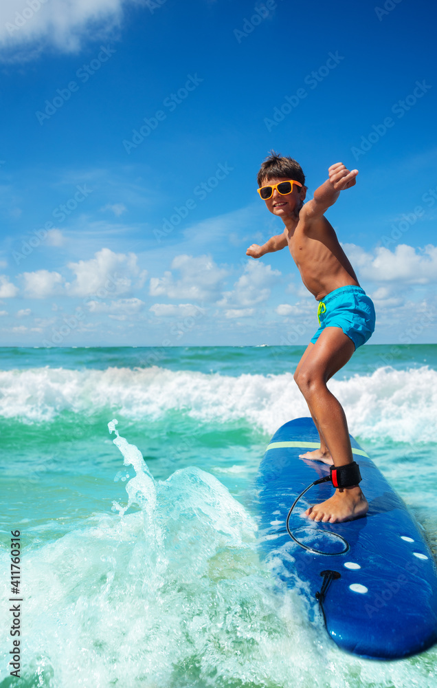 Photo of a boy riding surfboard on the small sea waves practicing with his board in cute sunglasses