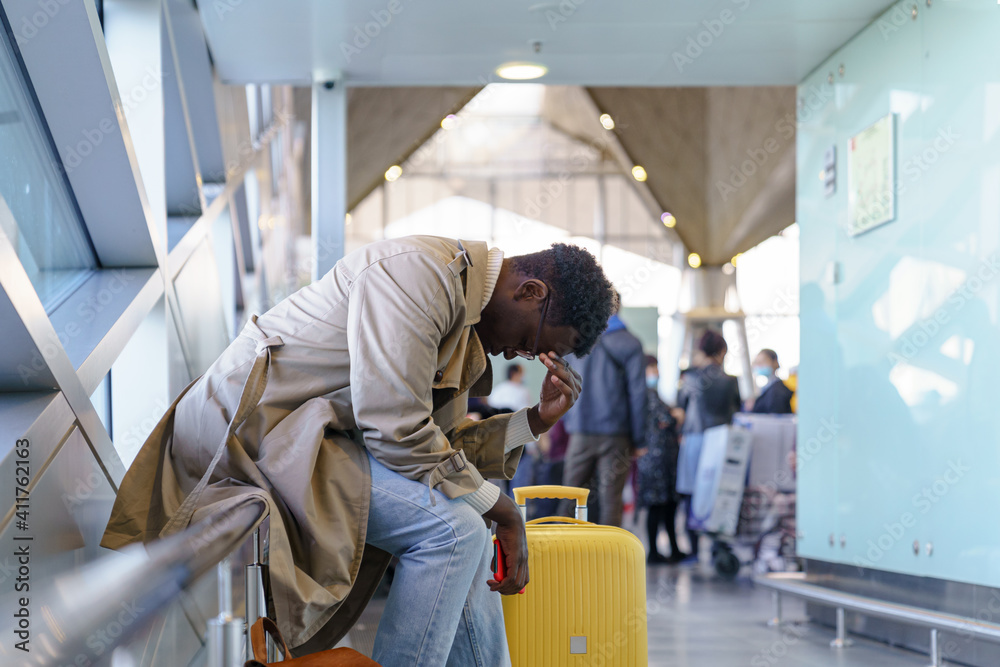 Black young man upset about the cancellation flight. Afro-American traveler male overslept his flight, missed the plane, touching her head, holding cellphone, sitting in airport terminal. Black young man upset about the cancellation flight. Afro-American traveler male overslept his flight, missed the plane, touching her head, holding cellphone, sitting in airport terminal.
