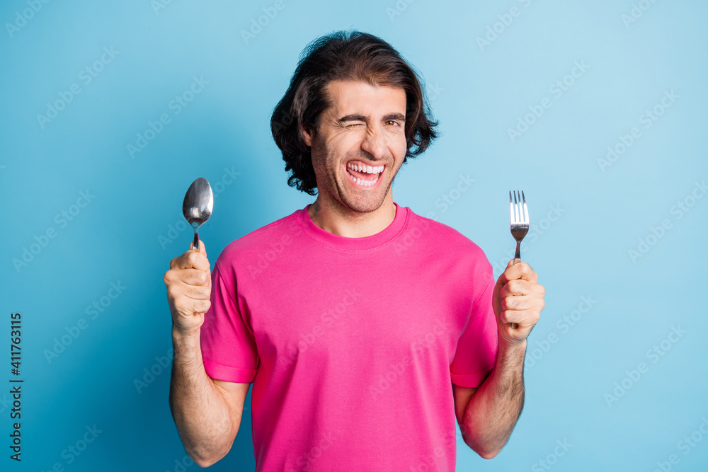 Portrait of cheerful guy holding in hands fork spoon winking healthy snack lunch expect isolated over bright blue color background Portrait of cheerful guy holding in hands fork spoon winking healthy snack lunch expect isolated over bright blue color background