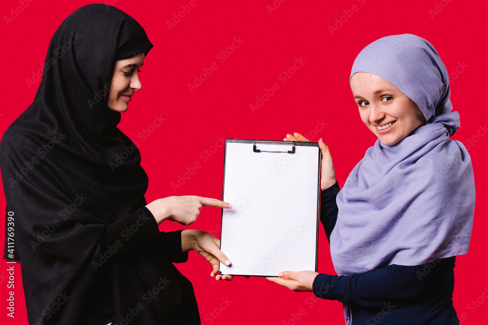 Smiling Muslim friends in hijab hold a folder in their hands, pointing with a forefinger to a white sheet. Education concept on red background. Smiling Muslim friends in hijab hold a folder in their hands, pointing with a forefinger to a white sheet. Education concept on red background.