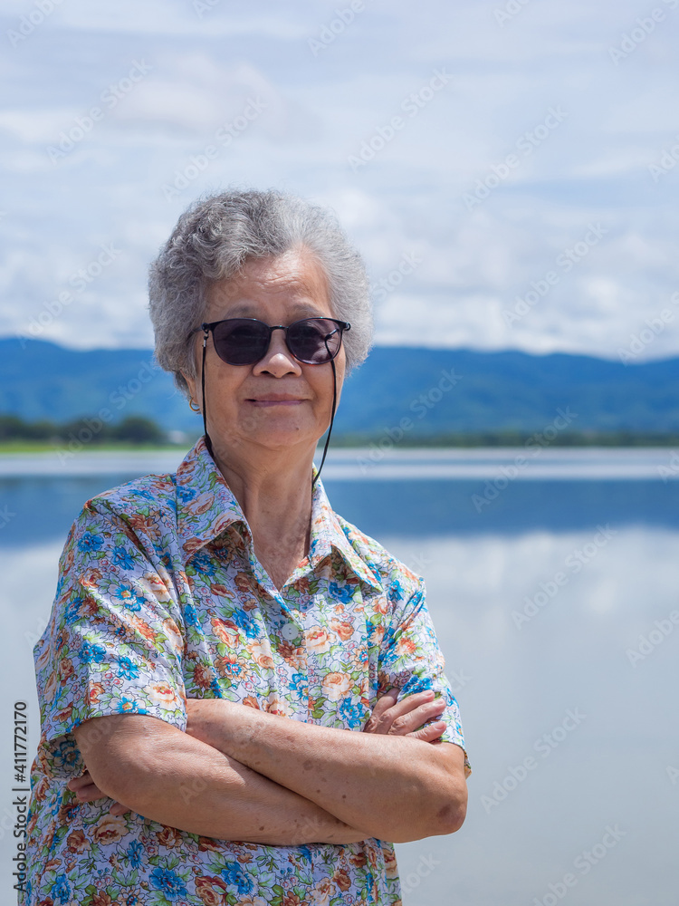 Portrait of senior woman wearing sunglasses arms crossed, smiling and looking at the camera while standing beside the lake with mountains backgrounds. Concept of Aged people and relaxation