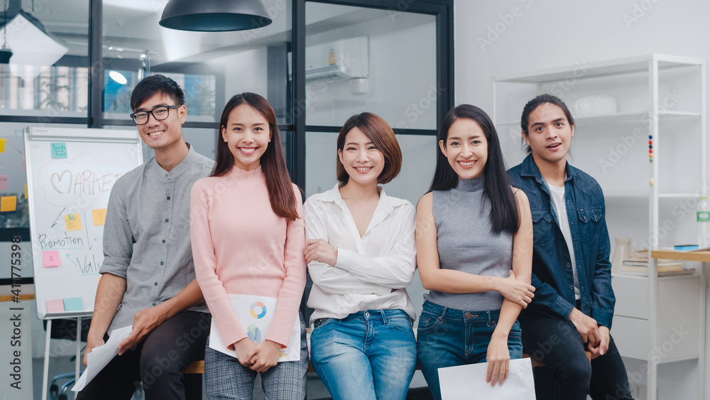 © tirachard - Group of Asia young creative people in smart casual wear looking at camera and smiling in creative office workplace. Diverse Asian male and female stand together at startup. Coworker teamwork concept.