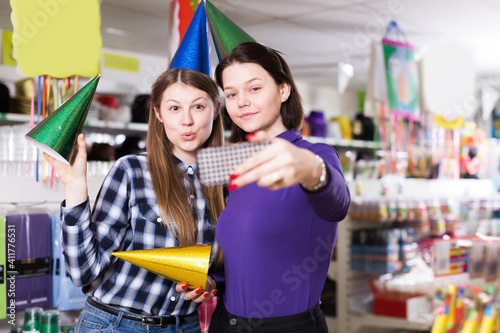 Comically dressed smiling girls making funny selfies photo in festive accessories shop..