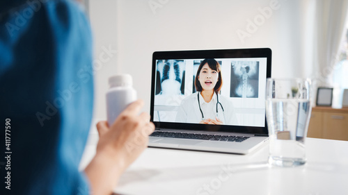 Young Asia lady using computer laptop talk about a disease in video conference call with senior doctor online consultation in living room at home. Social distancing, quarantine for coronavirus concept