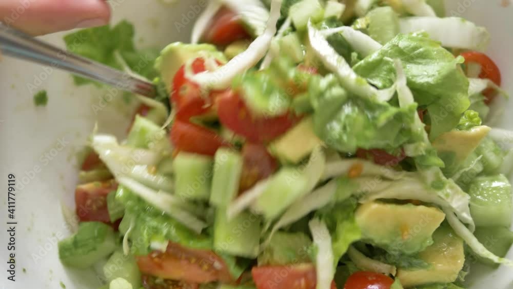 Woman mixes delicious fresh salad of lettuce avocado and small cherry tomatoes with spoon in white bowl close upper view Woman mixes delicious fresh salad of lettuce avocado and small cherry tomatoes with spoon in white bowl close upper view