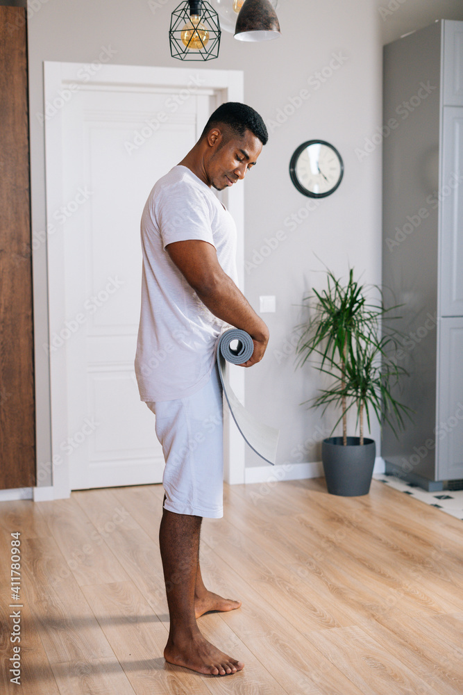 Side view of happy muscular African-American sportsman rolling yoga mat after finishing working out exercising at bright domestic room. Concept of sport training at home gym. Side view of happy muscular African-American sportsman rolling yoga mat after finishing working out exercising at bright domestic room. Concept of sport training at home gym.