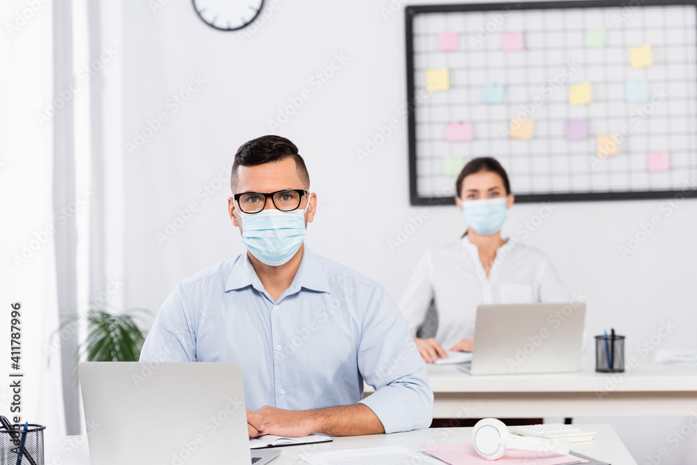 businessman in medical mask and glasses looking at camera near businesswoman on blurred background