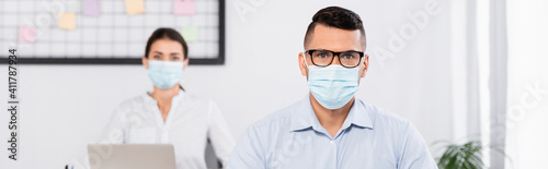 businessman in medical mask and glasses looking at camera near businesswoman on blurred background, banner