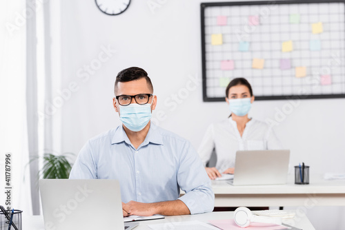businessman in medical mask and glasses looking at camera near businesswoman on blurred background