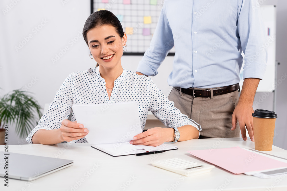 cheerful businesswoman looking at document near coworker standing at desk